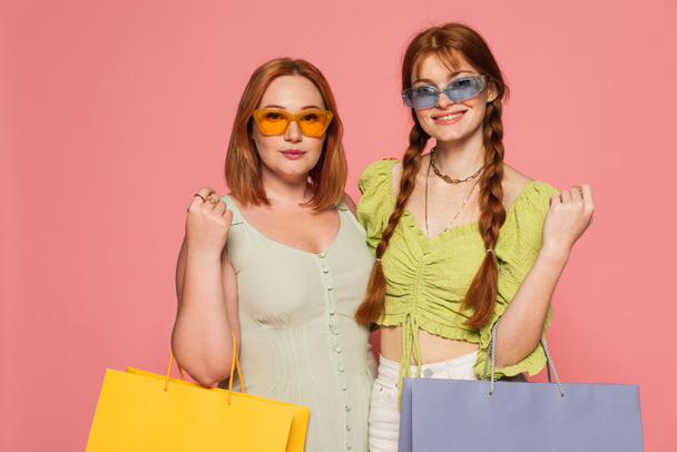 Κομψό σώμα θετικοί φίλοι σε γυαλιά ηλίου κρατώντας τσάντες ψώνια που απομονώνονται σε ροζ  - Φωτογραφία, εικόνα