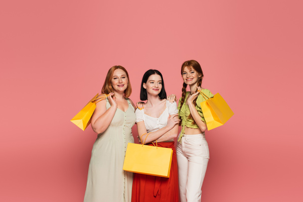 Young body positive women with shopping bags hugging isolated on pink  - Фото, изображение