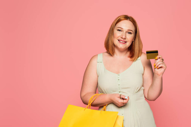 Positive plus size woman holding credit card and shopping bags isolated on pink  - Foto, Imagem