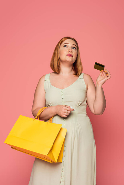 Pensive plus size woman with credit card and shopping bags isolated on pink  - Фото, зображення
