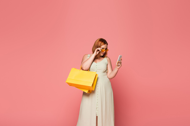 Stylish body positive woman holding shopping bags and smartphone isolated on pink  - 写真・画像