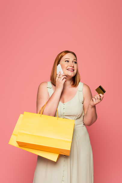Smiling plus size woman talking on smartphone and holding credit card and shopping bags isolated on pink  - Φωτογραφία, εικόνα