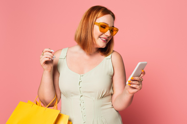 Smiling plus size woman in sunglasses using smartphone and holding shopping bags on pink background  - Photo, Image