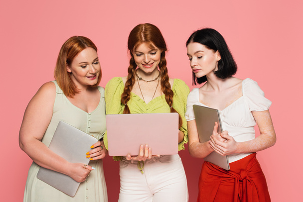 glimlachende vrouwen op zoek naar vriend met behulp van laptop op roze achtergrond  - Foto, afbeelding
