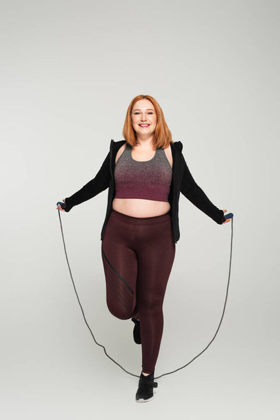 Cheerful body positive woman holding skipping rope on grey background - Фото, зображення