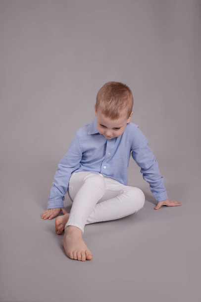 γοητευτικό μικρό παιδί προσχολικής ηλικίας σε λευκό παντελόνι και μπλε πουκάμισο σε γκρι φόντο. Παιδί, παιδί. - Φωτογραφία, εικόνα