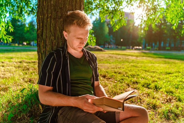 若い男が木の下に座って本を読んでいる。日没の緑の夏の公園 - 写真・画像