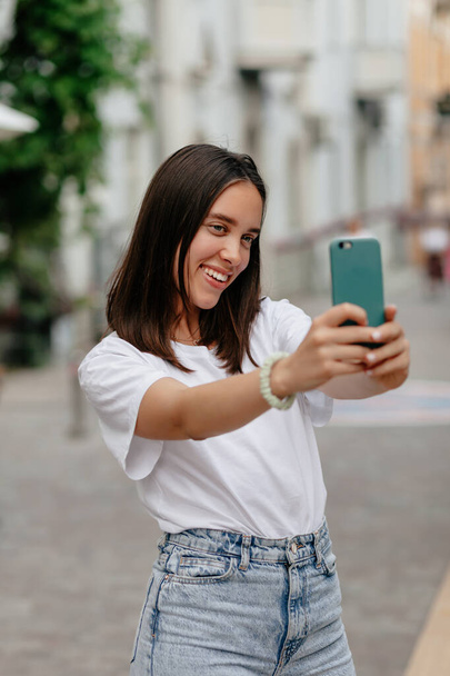 Charming adorable lady with short hairstyle wearing white t-shirt making selfie with smartphone while walking on the street . High quality photo - Photo, Image