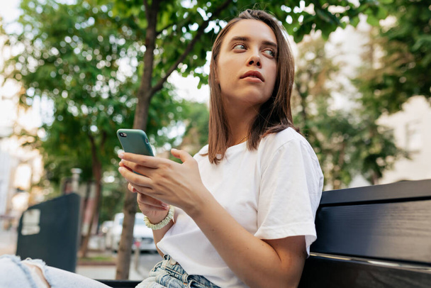 Close-up of stylish woman in white shirt against backdrop of city park. Lady is scrolling smartphone and looking away with interest  - Valokuva, kuva