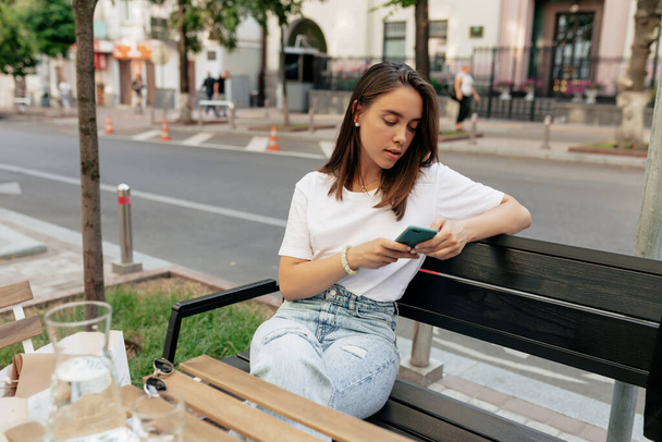 Pewna siebie nowoczesna kobieta z prostą fryzurą w białej koszuli i dżinsach siedząca na letnim tarasie i przewijająca smartfon. Wysokiej jakości zdjęcie - Zdjęcie, obraz