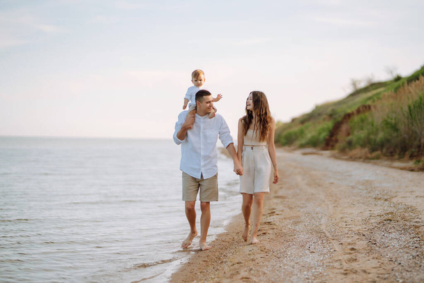 Cheerful young family with little baby boy spending time together on the beach. Father, mother and child against the background of the blue sea and sky. Family, childhood, active lifestyle concept. - Fotoğraf, Görsel