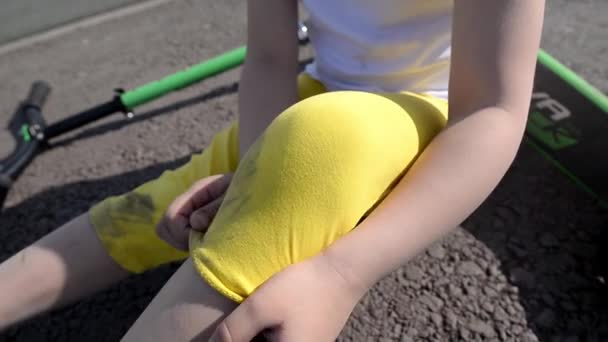 een kind in gele shorts vertoont een kleine schaafwond na het rijden in het park op de speelplaats op een 2-wielige scooter - Video