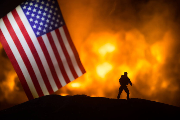 Amerykańska mała flaga na płonącym ciemnym tle. Koncepcja kryzysu wojny i konfliktów politycznych między narodami. Sylwetka uzbrojonego żołnierza przeciwko fladze USA. Skupienie selektywne - Zdjęcie, obraz