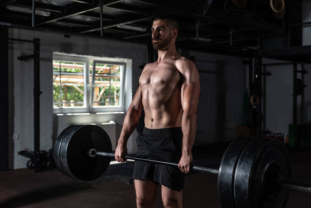 Young active sweaty strong muscular fit man with big muscles holding heavy barbell weight and doing hardcore cross weightlifting or deadlift workout training in the gym real people exercising - Фото, зображення