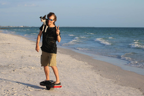 Vooraanzicht van de mens op hoverboard rijden met hond op schouder langs de oceaan voorkant - Foto, afbeelding