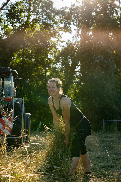 夕方の太陽の下で畑で干し草と遊ぶ魅力的な若い農家の女性。背景に森と赤いトラクター。ポジティブで幸せそうに見える女性にカメラ. - 写真・画像