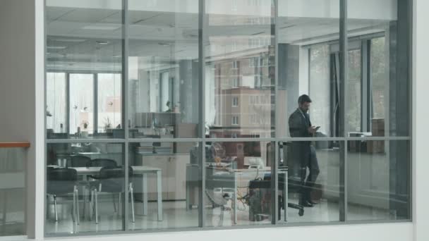 Door glazen muren slow mo shot van jonge mannelijke advocaat in zwart pak met behulp van smartphone tijdens het lopen langs moderne kantoor kamer in groot advocatenkantoor - Video