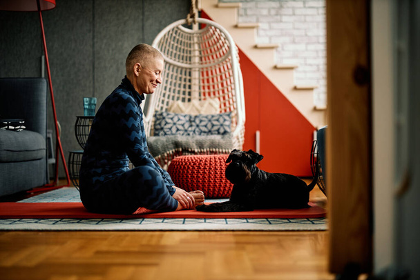 Χαρούμενη θετική ηλικιωμένη γυναίκα κάθεται στο πάτωμα στο σπίτι στο στρώμα γιόγκα και εξάσκηση γιόγκα με το σκυλί της. - Φωτογραφία, εικόνα
