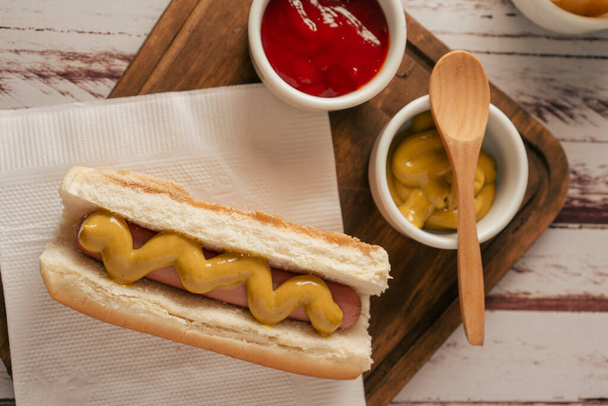 Vista superior de un perrito caliente con mostaza sobre una tabla de madera con macetas con ketchup y mostaza. Concepto de comida rápida. - Foto, imagen