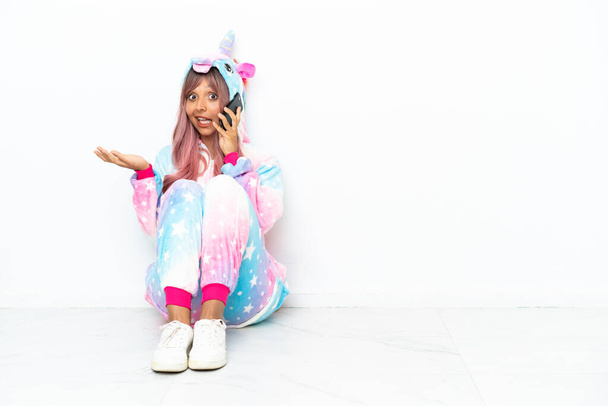 Giovane donna razza mista indossa un pigiama unicorno seduto sul pavimento isolato su sfondo bianco mantenendo una conversazione con il telefono cellulare con qualcuno - Foto, immagini