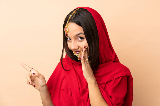 Young Indian woman isolated on beige background pointing to the side to present a product and whispering something - Foto, imagen