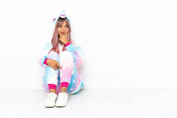 Young mixed race woman wearing a unicorn pajama sitting on the floor isolated on white background having doubts and with confuse face expression - Photo, image