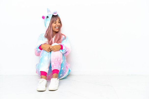 Joven mujer de raza mixta con un pijama unicornio sentado en el suelo aislado sobre fondo blanco riendo en posición lateral - Foto, imagen
