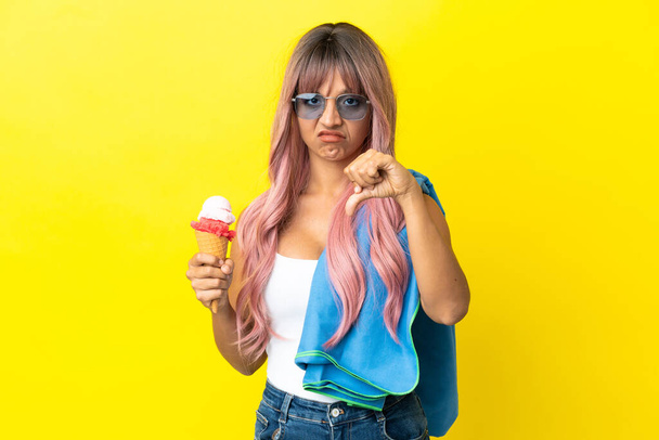 Молодая женщина смешанной расы с розовыми волосами с мороженым на желтом фоне показывает большой палец вниз с отрицательным выражением - Фото, изображение