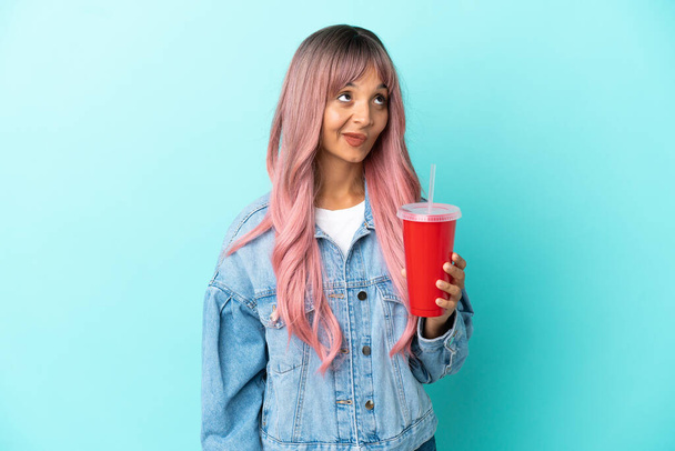 Young mixed race woman drinking a fresh drink isolated on blue background and looking up - Foto, immagini