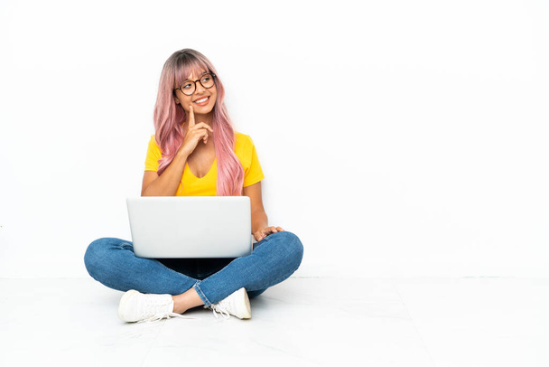 Junge gemischte Rasse Frau mit einem Laptop mit rosa Haaren sitzt auf dem Boden isoliert auf weißem Hintergrund und denkt eine Idee, während sie aufschaut - Foto, Bild