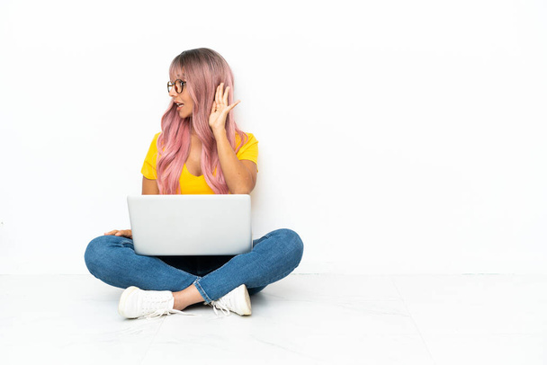 Молода змішана расова жінка з ноутбуком з рожевим волоссям, що сидить на підлозі ізольовано на білому тлі, слухаючи щось, поклавши руку на вухо
 - Фото, зображення