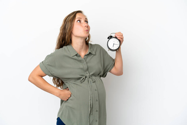 Молодая белая женщина на белом фоне, беременная и держащая часы, думая: - Фото, изображение