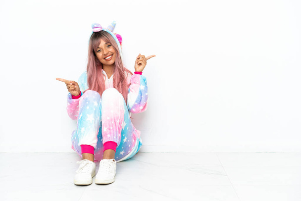 Giovane donna razza mista indossa un pigiama unicorno seduto sul pavimento isolato su sfondo bianco puntando il dito verso i laterali e felice - Foto, immagini
