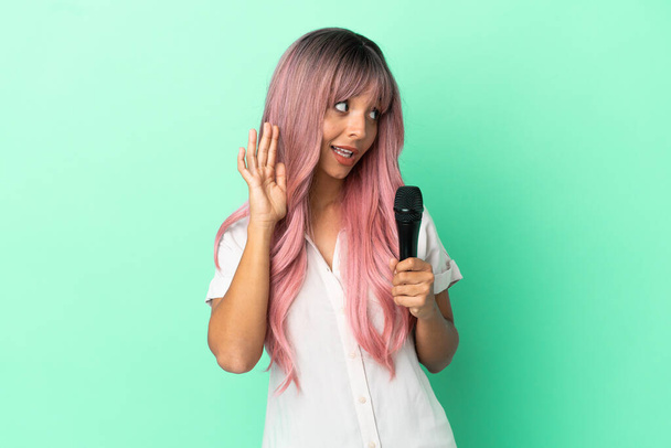 Молодая певица смешанной расы с розовыми волосами изолированы на зеленом фоне, слушая что-то, положив руку на ухо - Фото, изображение