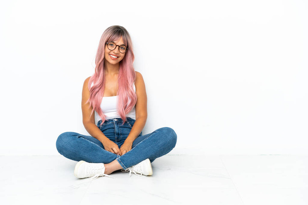 Молода змішана расова жінка з рожевим волоссям, що сидить на підлозі ізольовано на білому тлі сміється
 - Фото, зображення