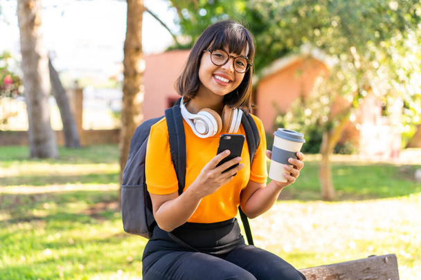 Νεαρή φοιτήτρια σε πάρκο που χρησιμοποιεί κινητό τηλέφωνο και κρατάει καφέ. - Φωτογραφία, εικόνα