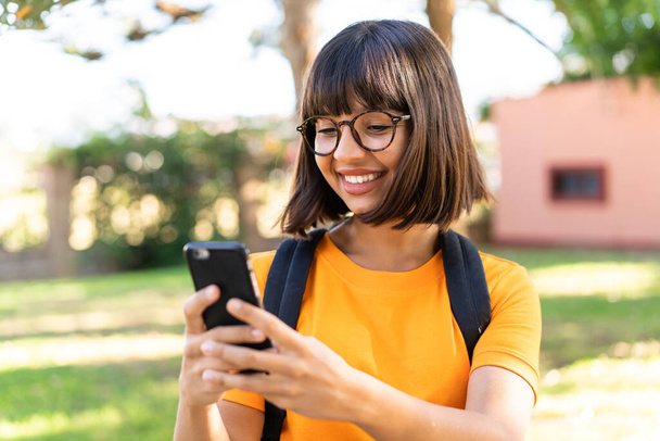 Молодая студентка в парке с помощью мобильного телефона - Фото, изображение