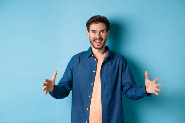 Glücklich lächelnder Mann zeigt großes Objekt, formt großes Ding und sieht erstaunt aus, steht auf blauem Hintergrund - Foto, Bild