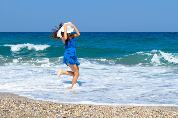 Happy beautiful carefree woman in blue dress, and white hat, walking barefoor on the beach. - Photo, Image
