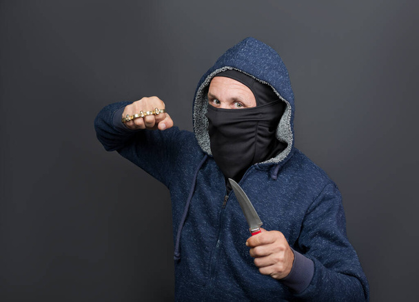 an offender attack with Brass knuckles and knife. Metal brass knuckles in the hand of American hooded man in mask. forbidden weapons in a fight, criminal. caucasian man strike fist with brass knuckles - 写真・画像