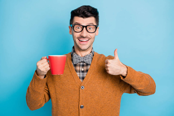 Photo of amazed brown haired man show thumb up hold cup wear plaid bow tie sweater isolated on blue color background - Foto, immagini