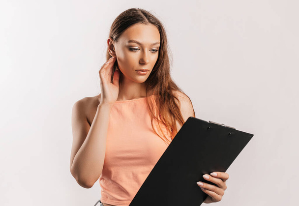 Girl student frowns with displeasure and reads information in documents. Beautiful young business woman holds a clipboard with mockup on gray background. Study and business concept. Online learning - Foto, imagen