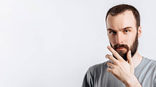 Serious puzzled handsome male model with beard holding hand on chin as if thinking about something, squinting at camera with suspicious look and standing over gray background. Man decides what to buy - Photo, image