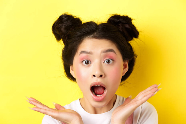 Close-up de mulher asiática animado com maquiagem glamour e penteado, gritando de espanto, boca aberta e olhar para a câmera em temor, fundo amarelo - Foto, Imagem