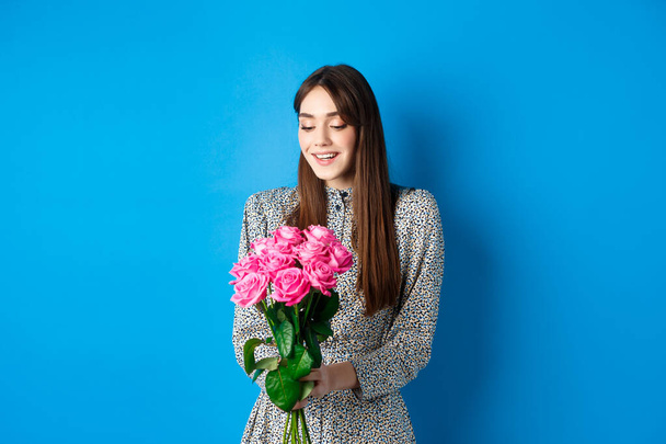 Valentines day concept. Romantic girl in dress looking happy at flowers, smiling at bouquet of pink roses, standing on blue background - Фото, зображення