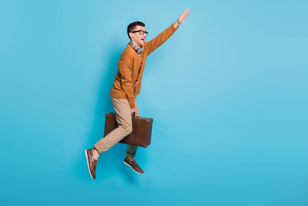 Full body profile side photo of brunette man ride suitcase jump air copyspace wear pants raise hand isolated on blue color background - Fotoğraf, Görsel