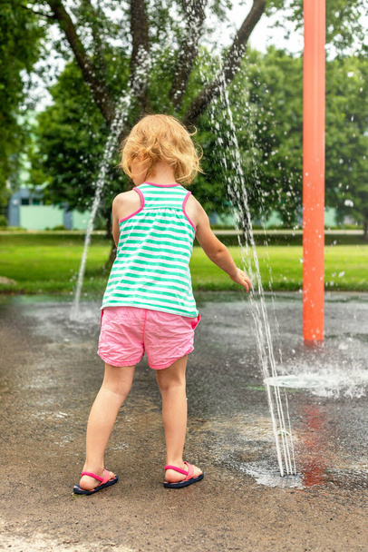 Küçük çocuk sıcak yaz gününde yerel halk parkında su sıçratma yerinde suyla oynuyor. Küçük güzel kız çeşme bahçesinde eğleniyor.. - Fotoğraf, Görsel