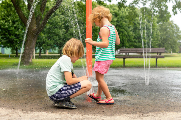 Little happy children playing with water at splash pad in the local public park on hot sunny summer day. Small beautiful kids having fun at fountain playground. - Photo, Image