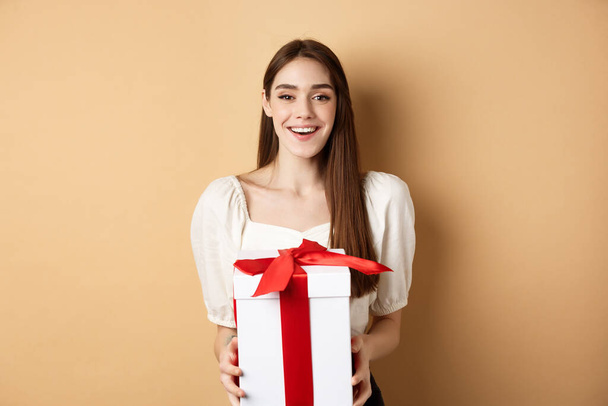 Happy valentines day. Cheerful smiling girl giving surprise gift to lover, looking at camera and holding present in box, standing on beige background - Photo, Image