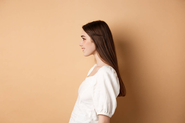 Profil pięknej białej kobiety z długimi naturalnymi włosami, patrząc w lewo na pustą przestrzeń i uśmiechnięty romantyczny, stojąc na beżowym tle - Zdjęcie, obraz